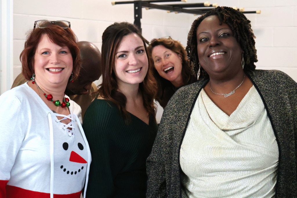 Four women smiling and posing together indoors, with festive outfits and joyful expressions, celebrating during a holiday gathering.