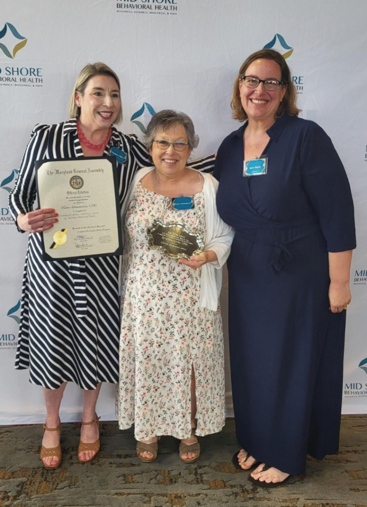 Three women smiling at an award ceremony, one holding a Maryland General Assembly citation and another holding a plaque, standing in front of a Mid Shore Behavioral Health backdrop.