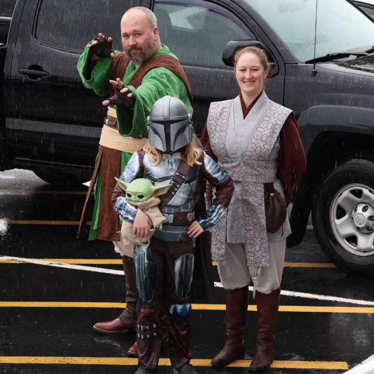 Three people dressed as Star Wars characters, including a Jedi, Mandalorian with a Grogu doll, and another Jedi, posing outdoors in the rain.