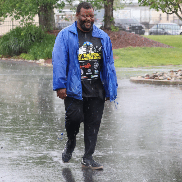 Smiling man walking in the rain wearing a blue jacket and black 5K event t-shirt with sponsor logos.