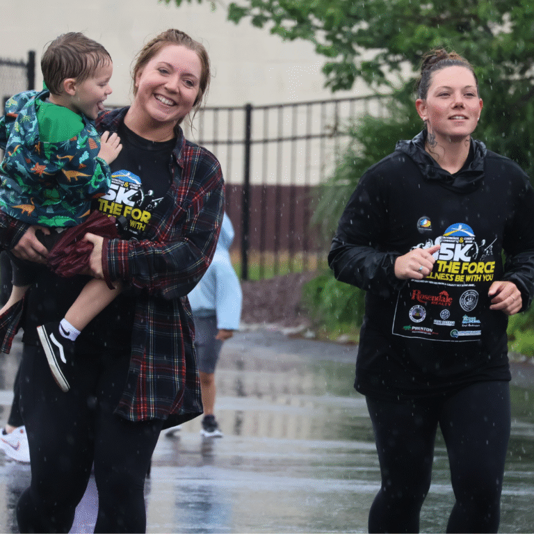 Two women at a 5K event in the rain, one carrying a smiling child and the other running in a black event hoodie.