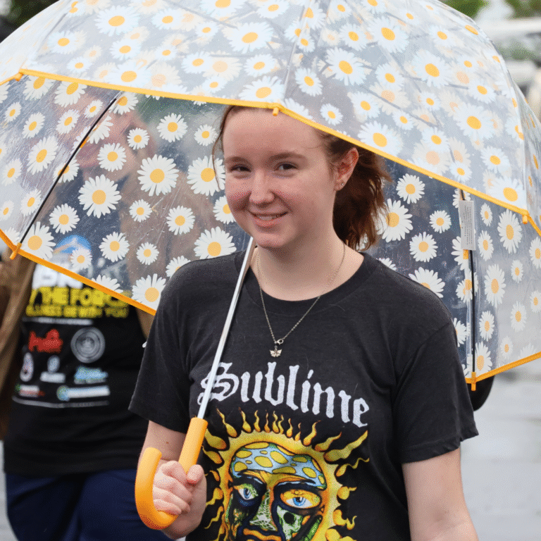Young girl smiling under a clear umbrella with white daisies during a rainy outdoor event.