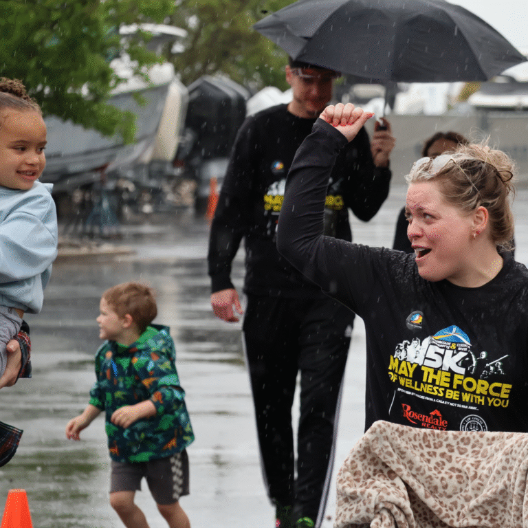 Woman in a black 5K event shirt raises her arm in celebration while looking at a smiling child being carried during a rainy outdoor event.