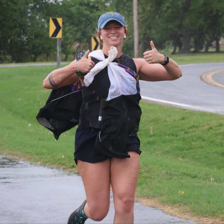 Woman running in the rain on a paved path, smiling and giving two thumbs up during a 5K event.