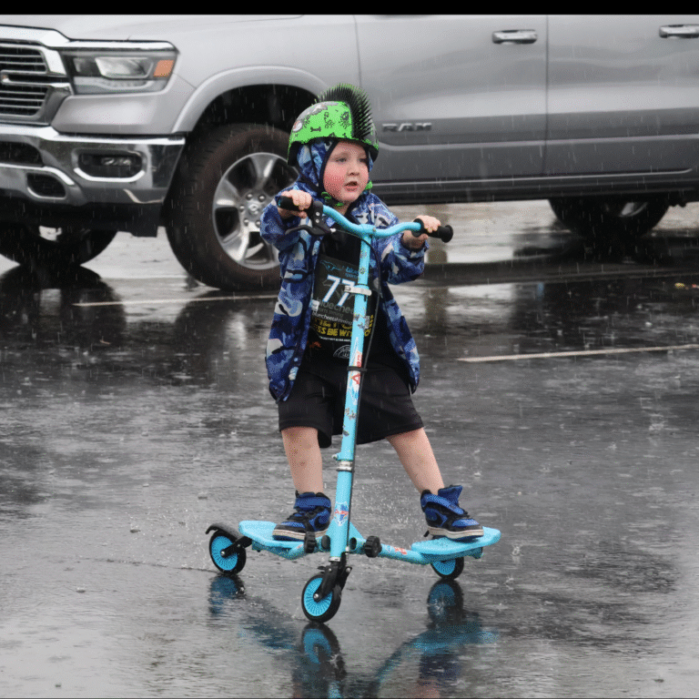 Young child riding a blue scooter in the rain, wearing a green spiked helmet and blue rain jacket at a 5K event.