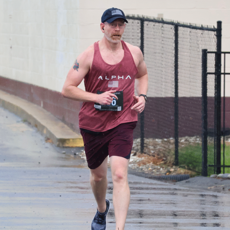 Man running in the rain during a 5K race, wearing a red tank top, maroon shorts, and a navy cap.