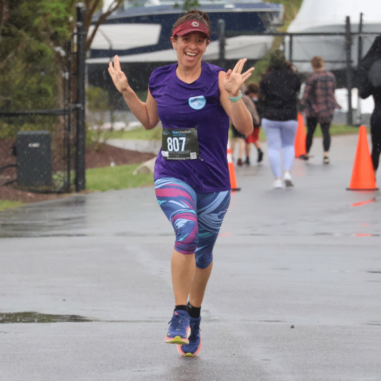 Smiling woman running in the rain during a 5K race, wearing a purple shirt, patterned leggings, and race bib number 807.