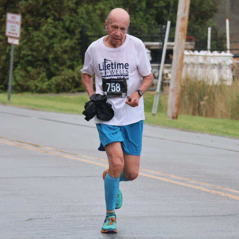 Older man running in the rain during a 5K event, wearing a white shirt, blue shorts, and race bib number 758.