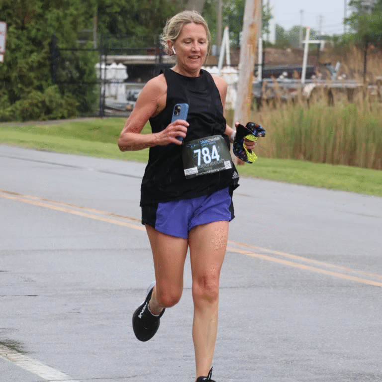 Woman running in the rain during a 5K, wearing a black tank top, purple shorts, and race bib number 784 while holding a phone and gear.