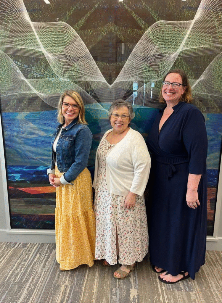 Three women smiling and standing in front of a colorful abstract wall mural, dressed in long dresses and casual attire.