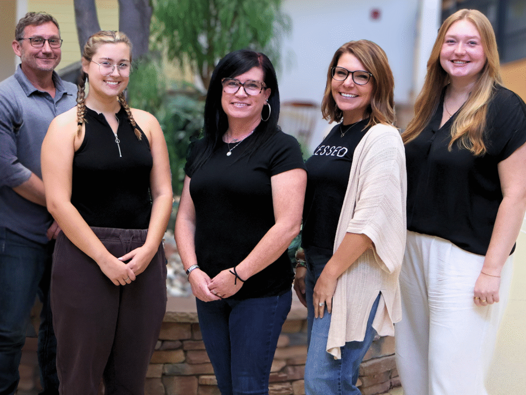 A group of five women and one man smiling indoors, representing a supportive workplace environment.